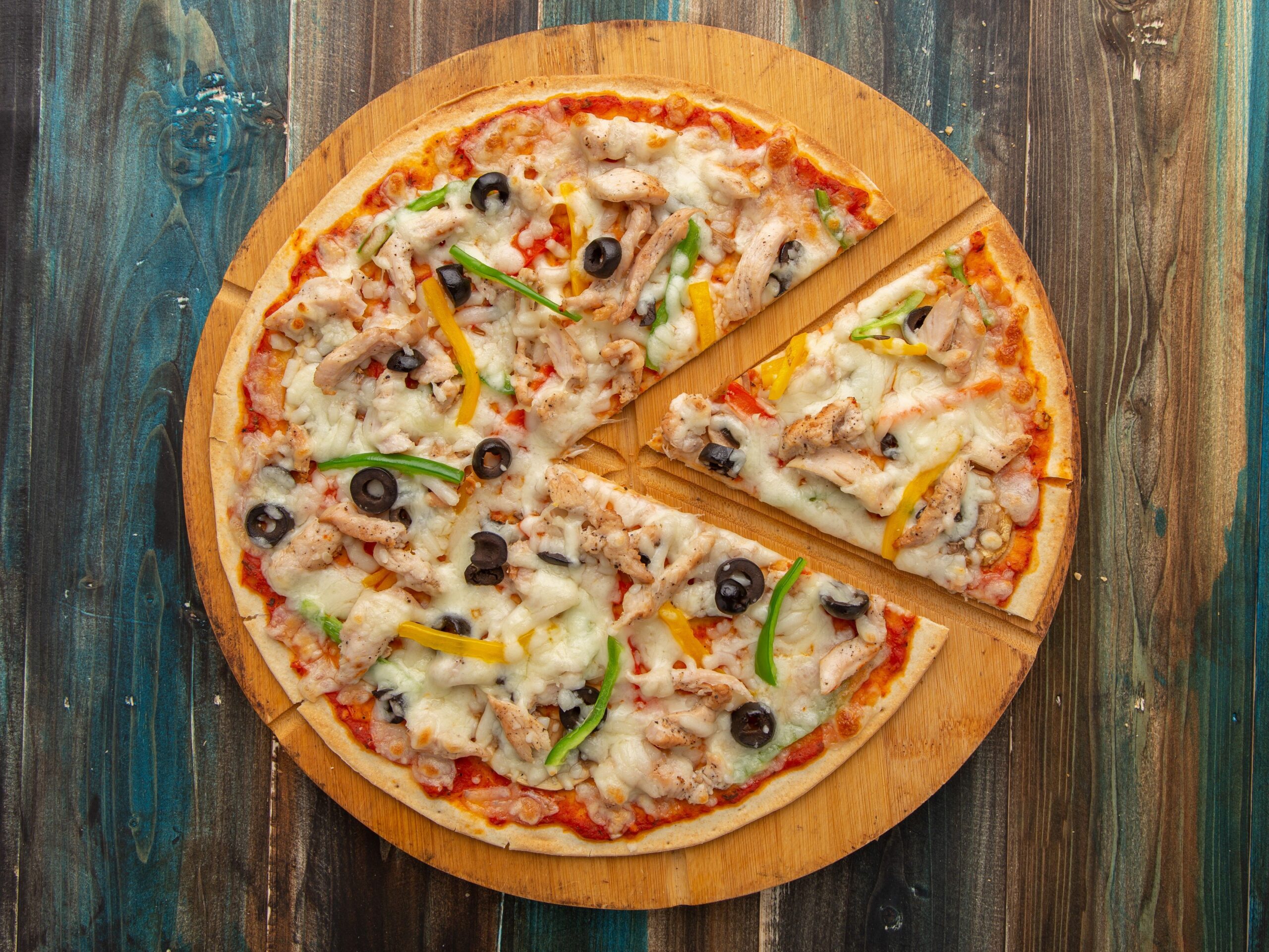 Top view of a chicken pizza with olives and bell peppers on a wooden board.